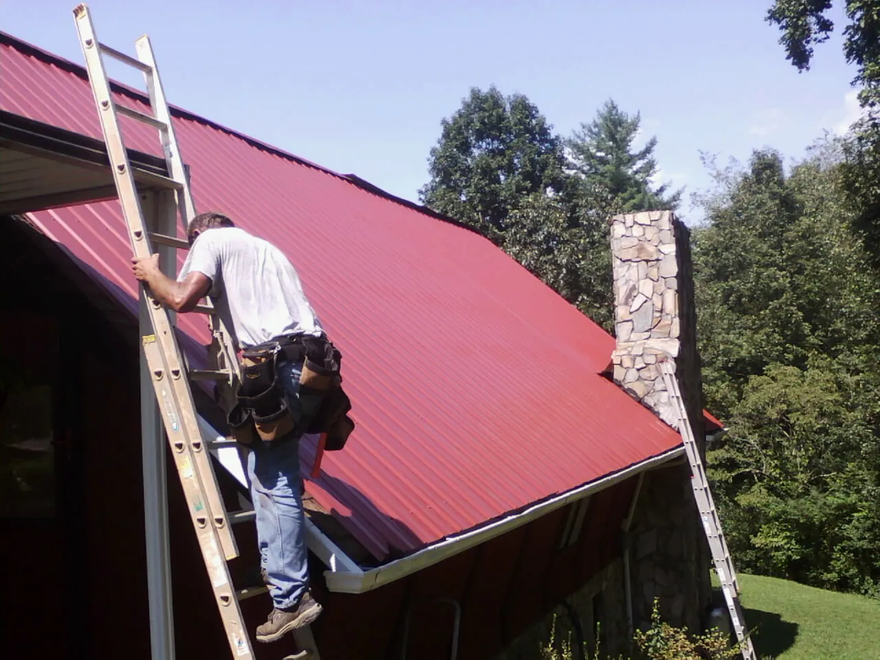 Roofer on a ladder beside a red metal roof and stone chimney — Soffit & Fascia Repair by Roofing Brothers in Juneau